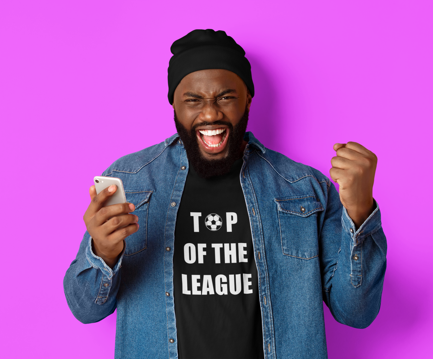 Man wearing a denim shirt and black beanie with 'Top of the League' T-Shirt Short Sleeve and a football graphic in the print, holding a phone on a purple background