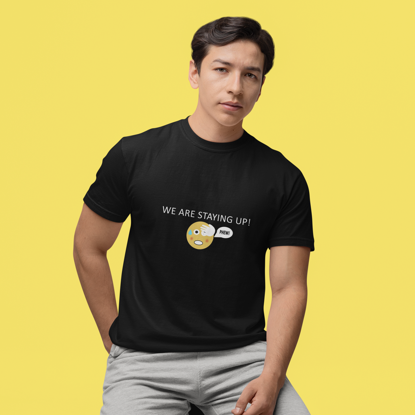 Man wearing a black short sleeve T-shirt with a round neck and the wording we are staying up with a phew emoji on a yellow studio background