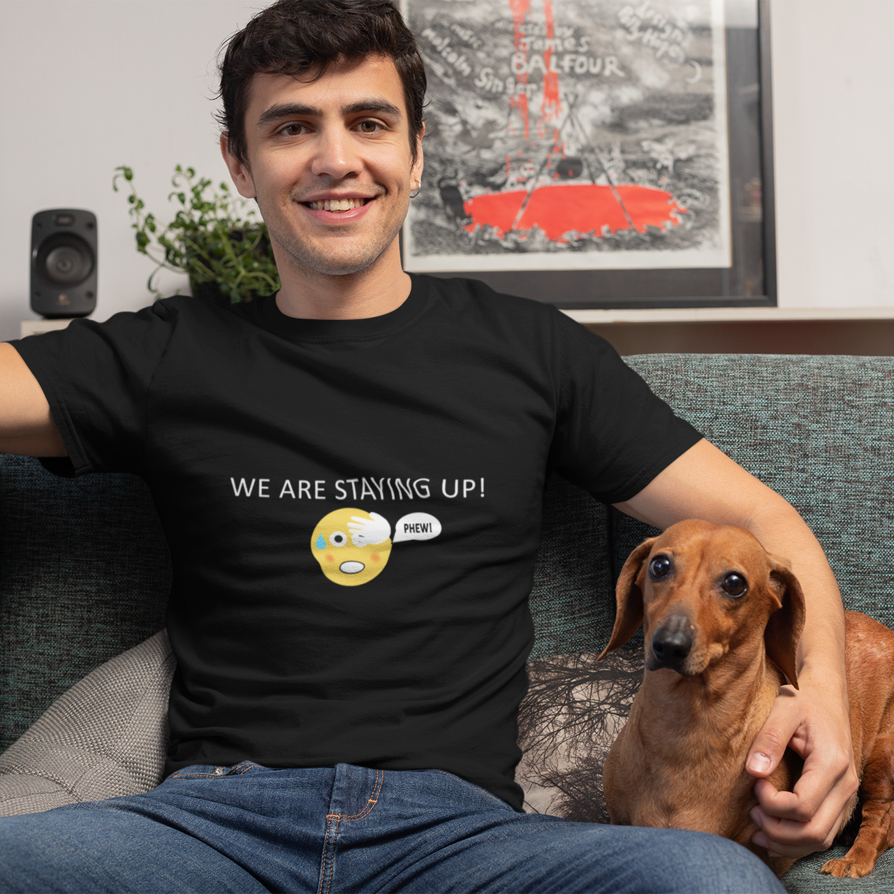 Man wearing a black t-shirt with text  of we are staying up with a emoji that shows the phew sign and a dog sitting on a couch in a living room.