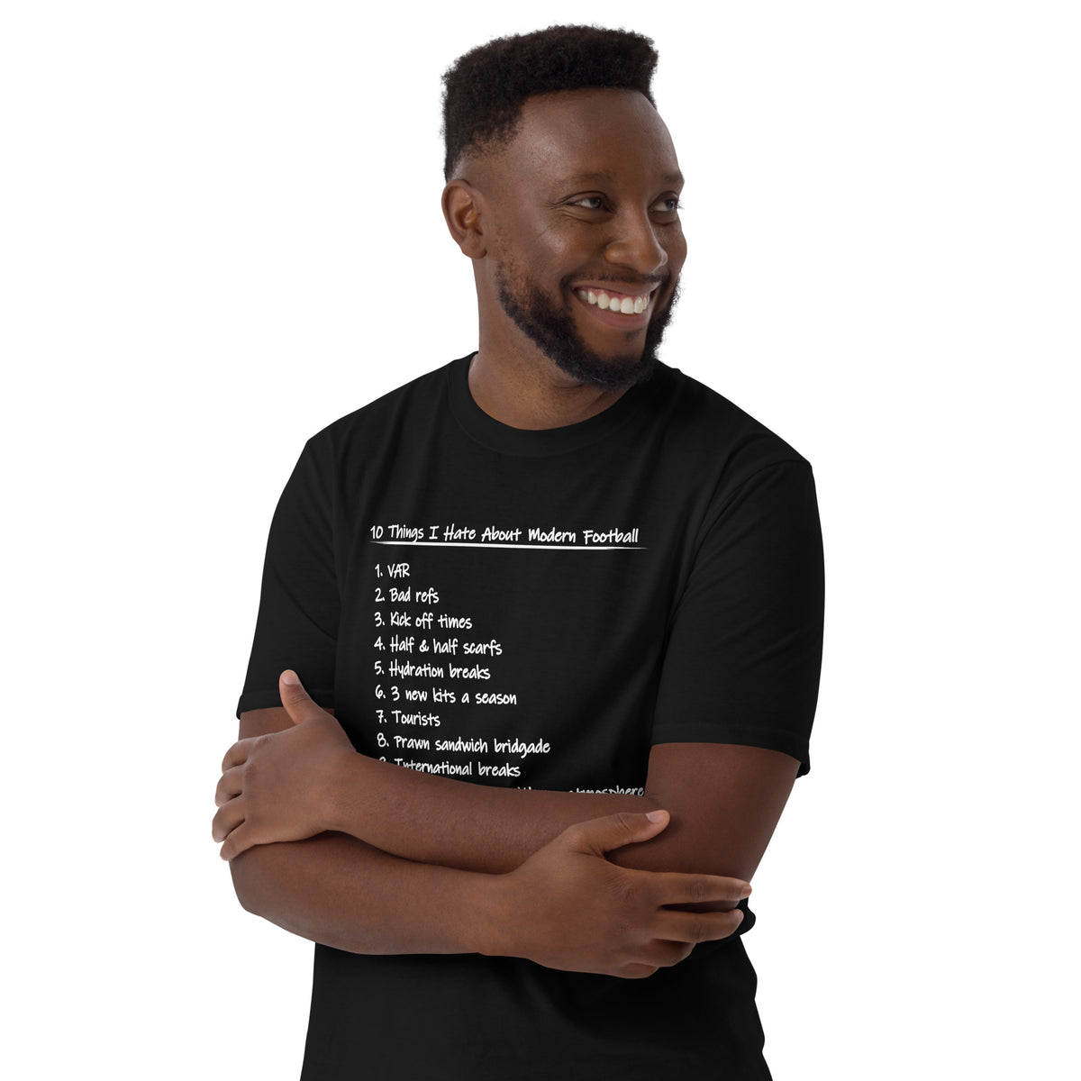 A smiling man wearing a black short-sleeve t-shirt with a white printed list of football-related phrases that people hate about modern football on the front, posed against a solid white background.