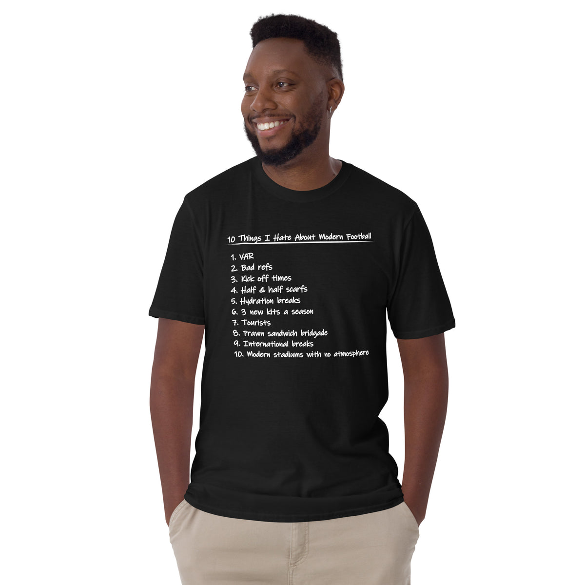 A smiling man wearing a black graphic t-shirt with a white printed list titled '10 Things I Hate About Modern Football' and light khaki pants, photographed against a solid white background.