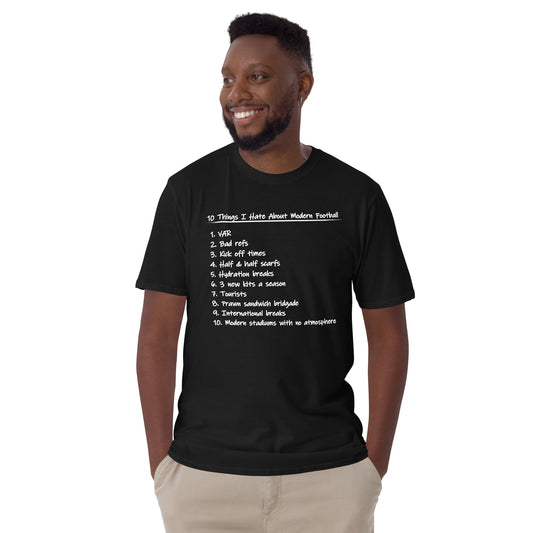 A smiling man wearing a black graphic t-shirt with a white printed list titled '10 Things I Hate About Modern Football' and light khaki pants, photographed against a solid white background.